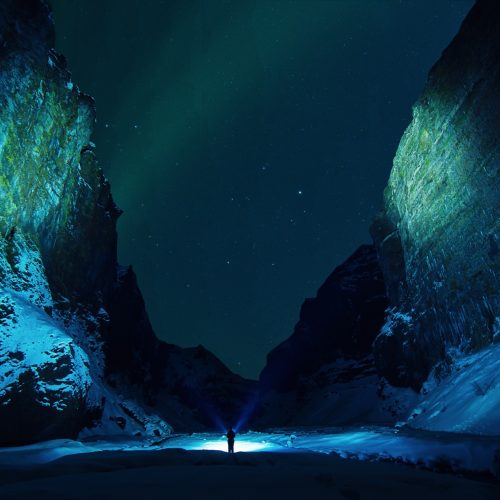 Person mit Lampe zwischen dunklen Felsen unter grünem Polarlicht